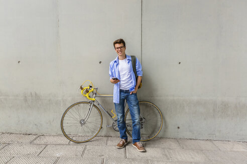 Lächelnder junger Mann mit Rennrad und Handy vor einer Betonwand - MGIF00151