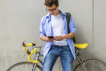 Junger Mann mit Rennrad, der vor einer Betonwand auf sein Handy schaut - MGIF00152
