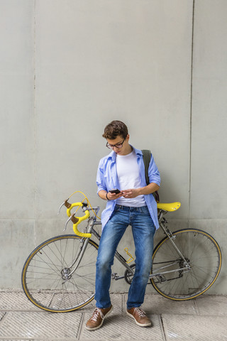 Junger Mann mit Rennrad, der vor einer Betonwand auf sein Handy schaut, lizenzfreies Stockfoto