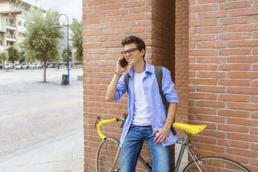 Porträt eines lächelnden jungen Mannes mit Rennrad am Telefon vor einer Backsteinmauer stehend - MGIF00157