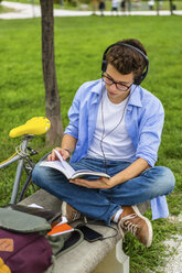 Junger Mann mit Rennrad und Kopfhörern sitzt auf einer Bank und liest ein Buch - MGIF00167
