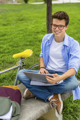 Porträt eines lächelnden jungen Mannes, der auf einer Bank sitzt und einen Laptop benutzt - MGIF00168