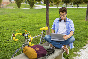 Junger Mann mit Rennrad auf einer Bank sitzend mit Laptop - MGIF00170