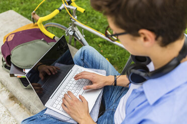 Young man with racing cycle sitting on a bench using laptop, elavated view - MGIF00171