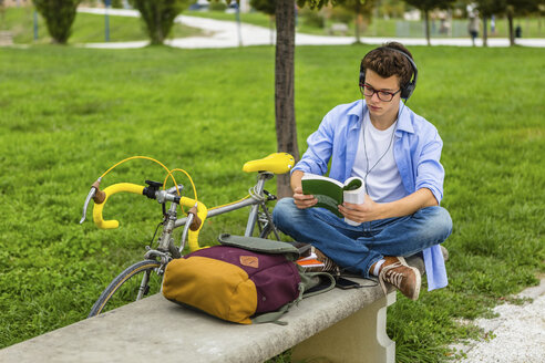 Young man with racing cycle and headphones sitting on a bench reading book - MGIF00172