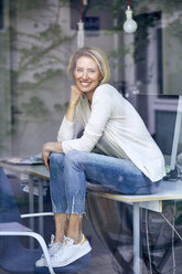 Porträt einer lachenden blonden Frau, die auf einem Schreibtisch in einem Büro sitzt und aus dem Fenster schaut - PNEF00010