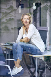 Portrait of blond woman sitting on desk in an office looking through window - PNEF00011