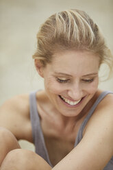 Porträt einer lachenden blonden Frau, die am Strand sitzt - PNEF00047