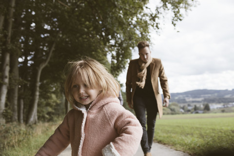 Porträt eines blonden kleinen Mädchens mit einem Vater, der ihr im Hintergrund nachläuft, lizenzfreies Stockfoto