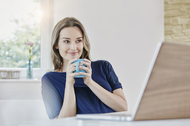 Smiling woman at home with coffee mug and laptop - RORF01017