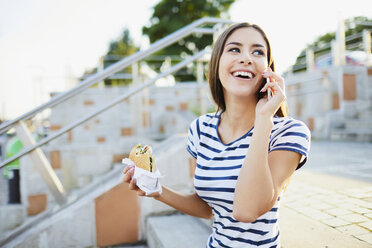 Junge Frau spricht am Telefon und isst Bagel in der Stadt - BSZF00067