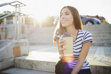 Junge Frau sitzt auf einer Treppe im Freien und hält Kaffee - BSZF00068