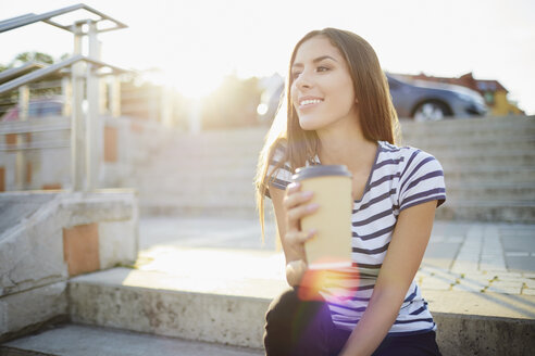 Young woman sitting on stairs outdoors and holding coffee - BSZF00068