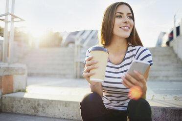 Young woman sitting on stairs in the city with phone and coffee - BSZF00069