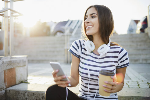 Woman with headphones sitting on stairs in the city with smartphone and coffee - BSZF00071