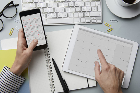 Draufsicht auf eine Frau, die ein Smartphone und ein Tablet mit Kalender auf dem Schreibtisch hält, lizenzfreies Stockfoto