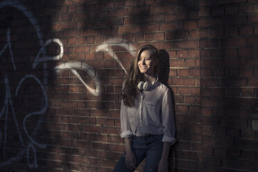 Portrait of laughing woman with headphones leaning against brick wall - PNEF00122