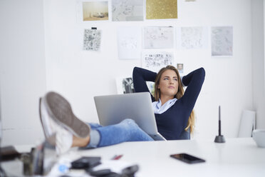 Junge Frau mit Laptop am Schreibtisch im Büro lehnt sich zurück - PNEF00141