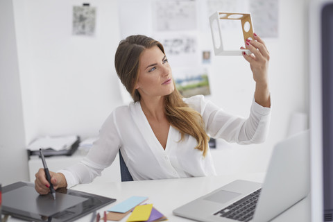 Junge Frau am Schreibtisch im Büro, die ein Modell hält und ein Grafiktablett benutzt, lizenzfreies Stockfoto