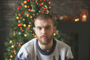 Porträt eines ernsten jungen Mannes mit Vollbart vor einem beleuchteten Weihnachtsbaum zu Hause - RTBF01050