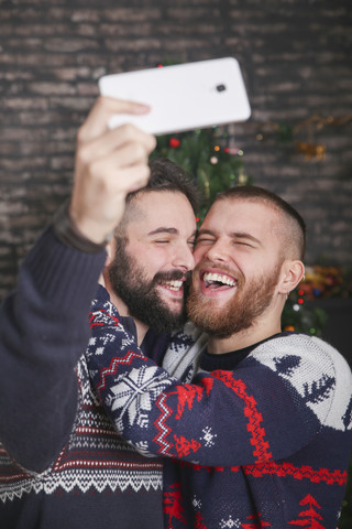 Porträt eines lachenden schwulen Paares, das zu Weihnachten zu Hause ein Selfie mit dem Smartphone macht, lizenzfreies Stockfoto