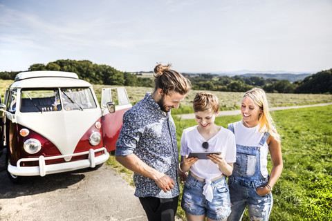 Glückliche Freunde vor einem Lieferwagen in einer ländlichen Landschaft, die auf ein Tablet schauen, lizenzfreies Stockfoto