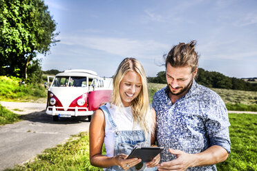 Glückliches Paar vor einem Lieferwagen in ländlicher Landschaft mit Blick auf ein Tablet - FMKF04546