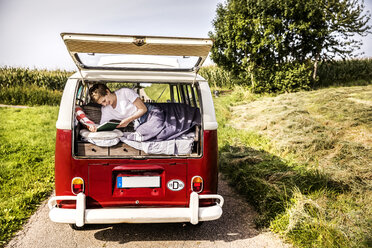 Happy woman lying in a van in rural landscape reading book - FMKF04566