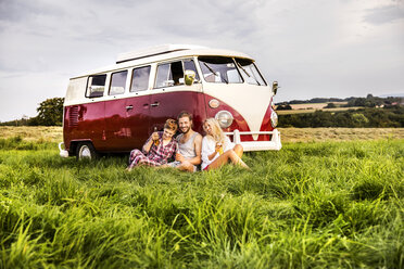 Happy friends with beer bottles sitting at a van in rural landscape - FMKF04604