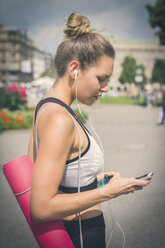 Junge Frau mit Yogamatte, Mobiltelefon und Kopfhörern in der Stadt - JUNF00931