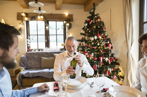 Lächelnder älterer Mann mit Familie, der eine Flasche Wein am Weihnachtstisch hält - HAPF02180