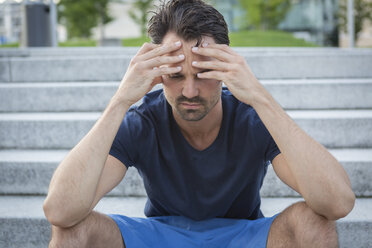 Athlete sitting on stairs with hands on head, having a headache - JUNF00976