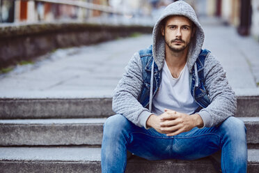 Serious man in hooded jacket sitting on stairs - BSZF00093