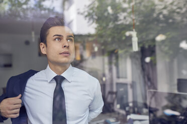 Porträt eines jungen Geschäftsmannes hinter einer Glasscheibe in einem Büro, der seine Jacke anzieht - PNEF00179
