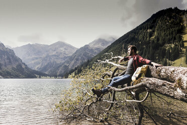 Austria, Tyrol, Alps, hiker relaxing on tree trunk at mountain lake - UUF11992