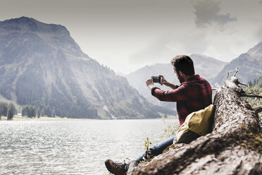 Austria, Tyrol, Alps, hiker sitting on tree trunk at mountain lake taking cell phone picture - UUF11993