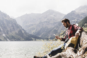 Austria, Tyrol, Alps, hiker relaxing on tree trunk at mountain lake checking cell phone - UUF11994