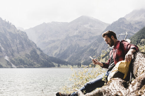 Österreich, Tirol, Alpen, Wanderer ruht sich auf einem Baumstamm am Bergsee aus und kontrolliert sein Handy - UUF11994