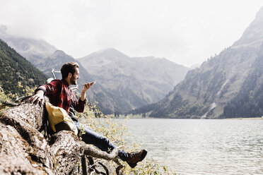 Austria, Tyrol, Alps, hiker on tree trunk at mountain lake using cell phone - UUF11995