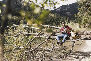 Österreich, Tirol, Alpen, Wanderer ruht sich auf einem Baumstamm am Bergsee aus und kontrolliert sein Handy - UUF11996