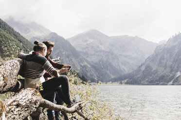 Österreich, Tirol, Alpen, Paar entspannt sich auf Baumstamm an Bergsee und benutzt Handy - UUF11997