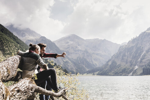 Österreich, Tirol, Alpen, Paar sitzt auf Baumstamm am Bergsee und macht Selfie - UUF11998