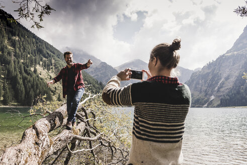 Österreich, Tirol, Alpen, Frau fotografiert mit dem Handy einen Mann, der auf einem Baumstamm an einem Bergsee balanciert - UUF11999