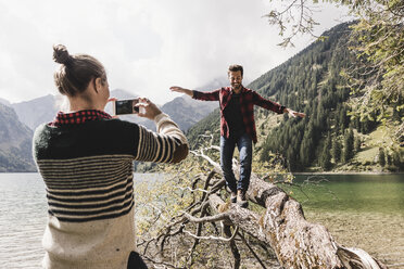 Österreich, Tirol, Alpen, Frau fotografiert mit dem Handy einen Mann, der auf einem Baumstamm an einem Bergsee balanciert - UUF12000