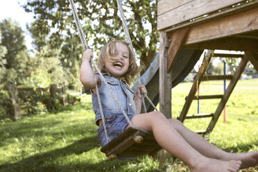 Laughing little girl sitting on swing in the garden - KMKF00023