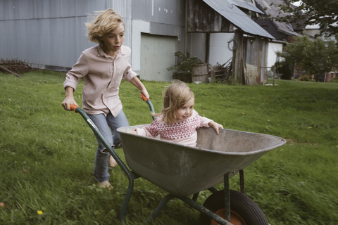 Junge schiebt Schubkarre mit seiner kleinen Schwester durch den Garten, lizenzfreies Stockfoto