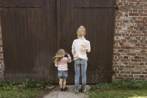 Rückansicht des Bruders und seiner kleinen Schwester vor einer Holztür, lizenzfreies Stockfoto