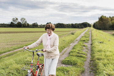 Ältere Frau schiebt Fahrrad in ländlicher Landschaft - UUF12024