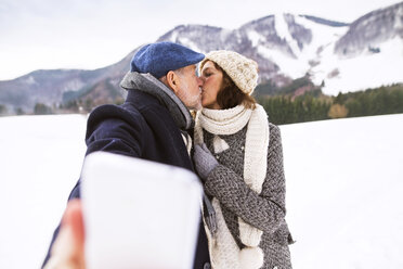 Älteres Paar, das sich in einer Winterlandschaft küsst, während es ein Selfie mit dem Handy macht - HAPF02235