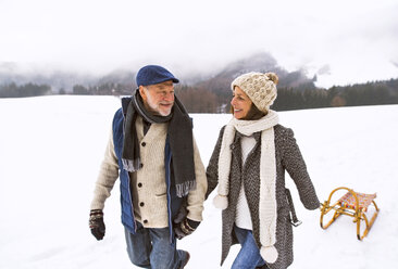 Happy senior couple with sledge walking in snow-covered landscape - HAPF02247